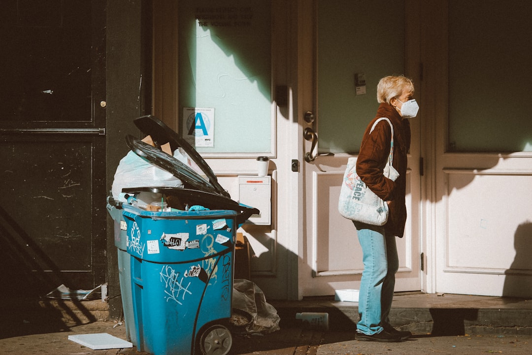 woman-in-brown-jacket-and-blue-denim-jeans-standing-beside-red-plastic-trash-bin-yolf8d1h-di